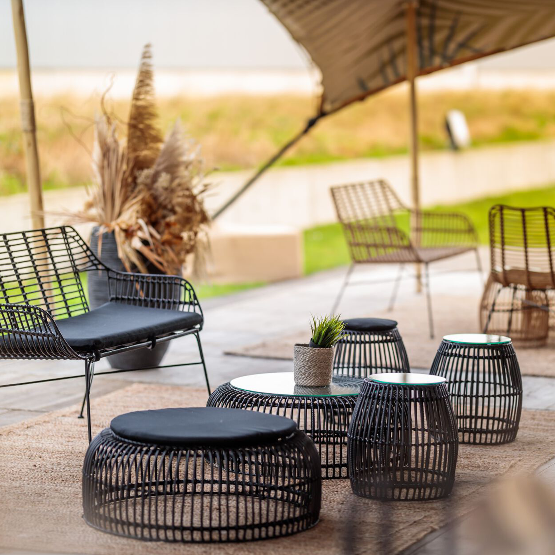 Outdoor seating area featuring a Palm Stacking Side Table - Black with glass top surrounded by matching black wireframe stools and a cushioned chair under a canopy.